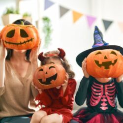 A mom and two daughters holding pumpkins up to their faces while wearing halloween costumes
