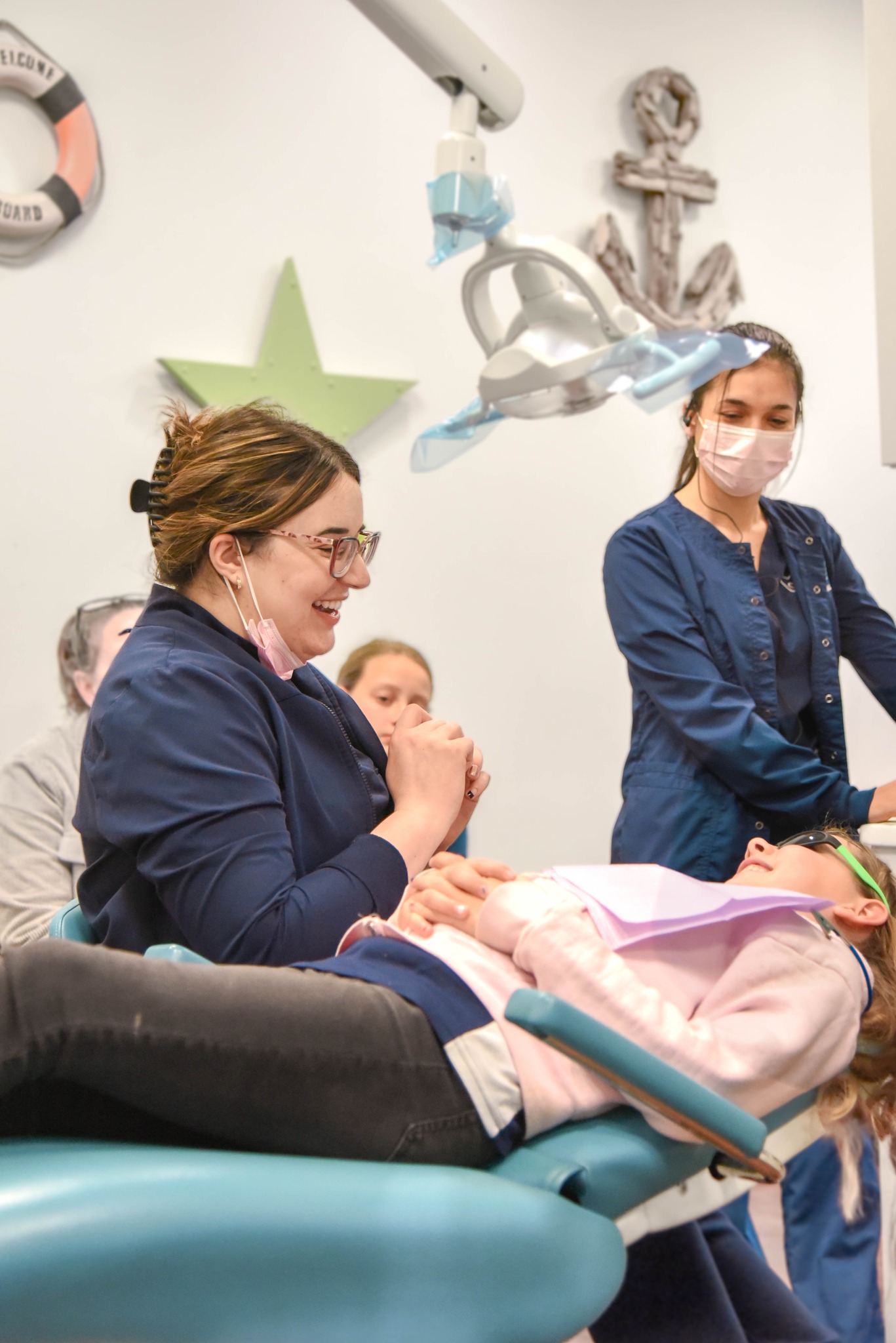 A pediatric dentist holds a young patient's hand and smiles during a dental checkup in a nautical-themed treatment room at Richmond Pediatric Dentistry and Orthodontics