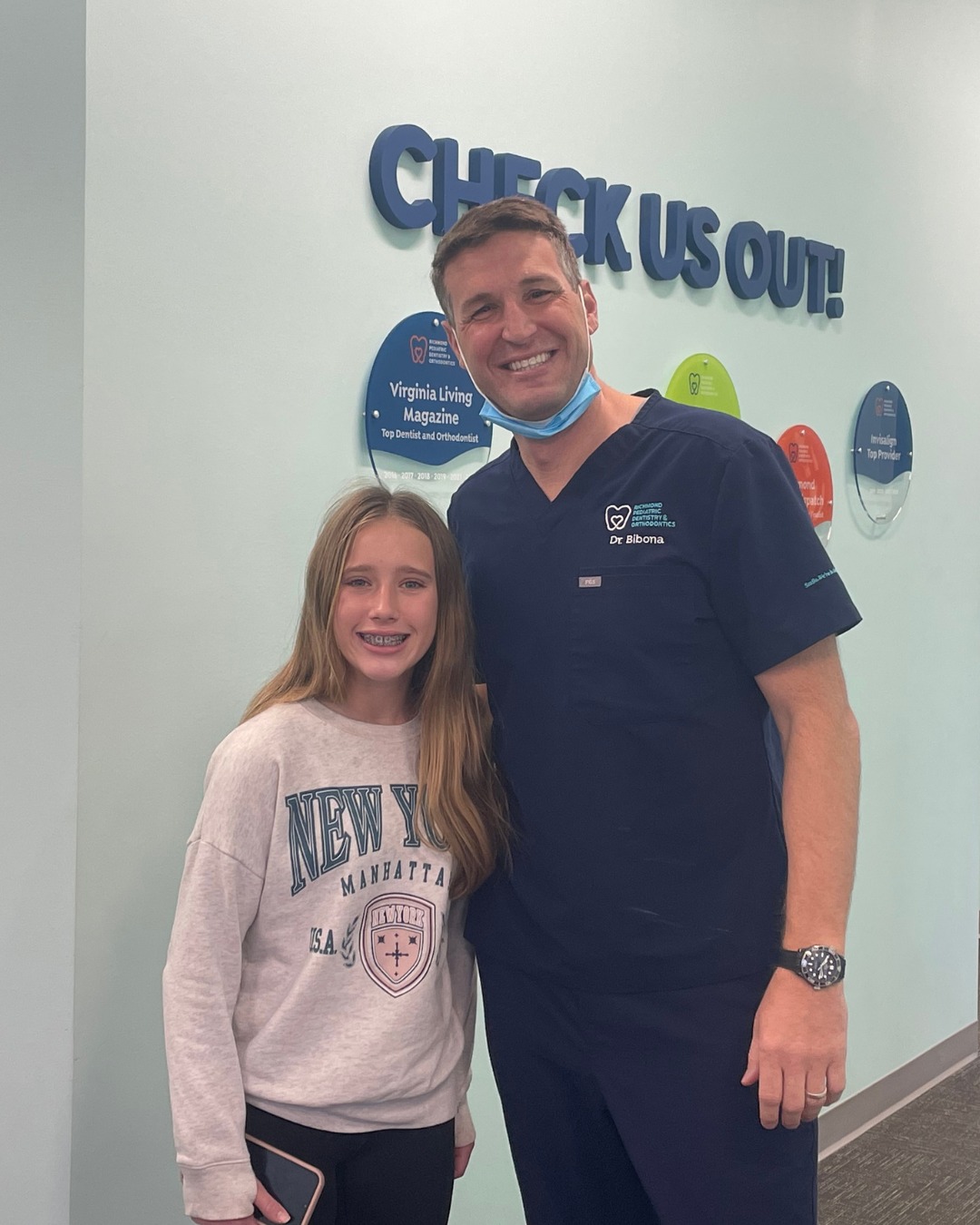 Dr. Kevin Bibona, orthodontist at Richmond Pediatric Dentistry and Orthodontics, smiling with a young braces patient in front of the office award wall featuring Virginia Living Magazine Top Dentist and Orthodontist recognition
