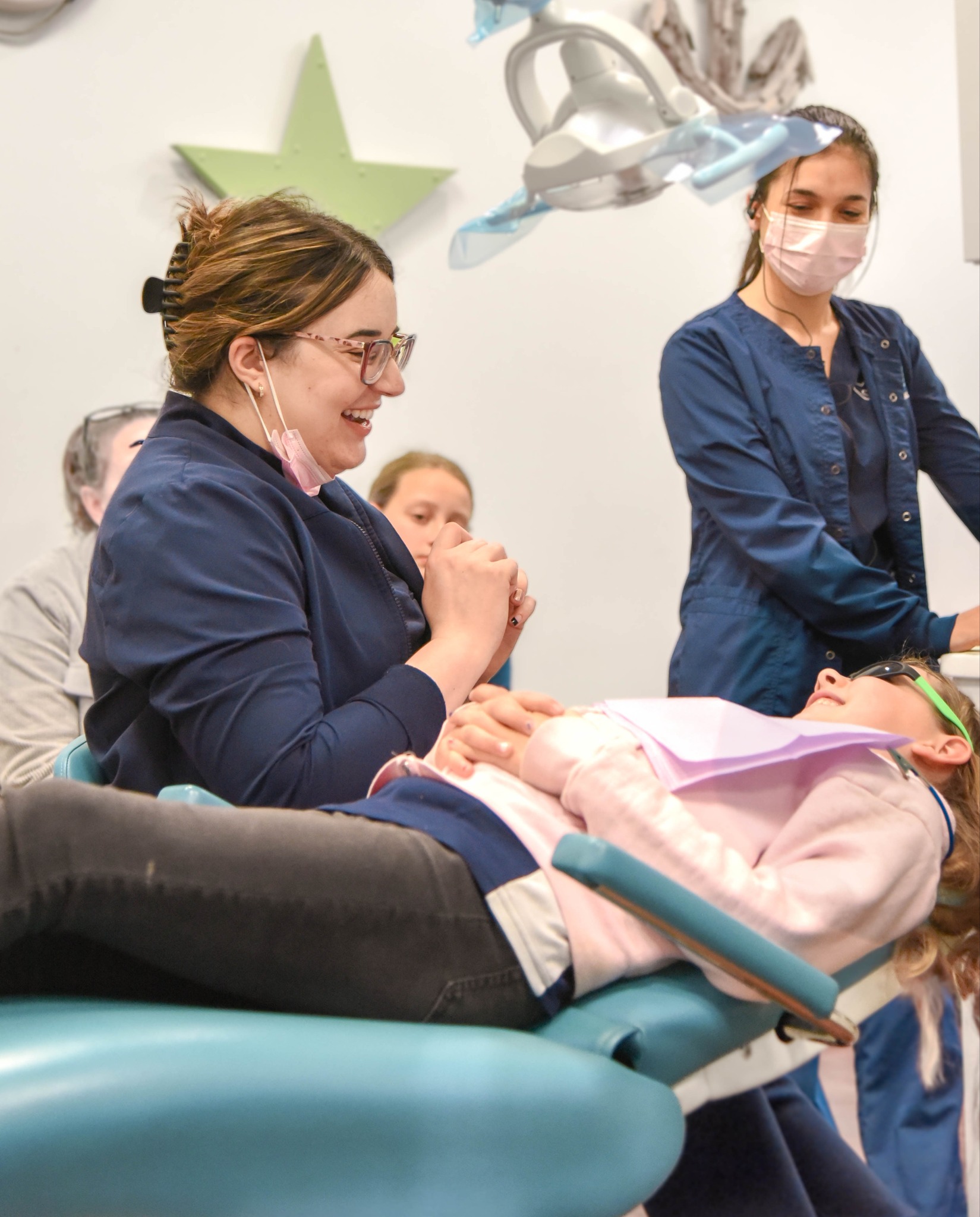 A pediatric dentist laughs with a young girl in a dental chair at an RPDO office, with a dental assistant assisting in the background
