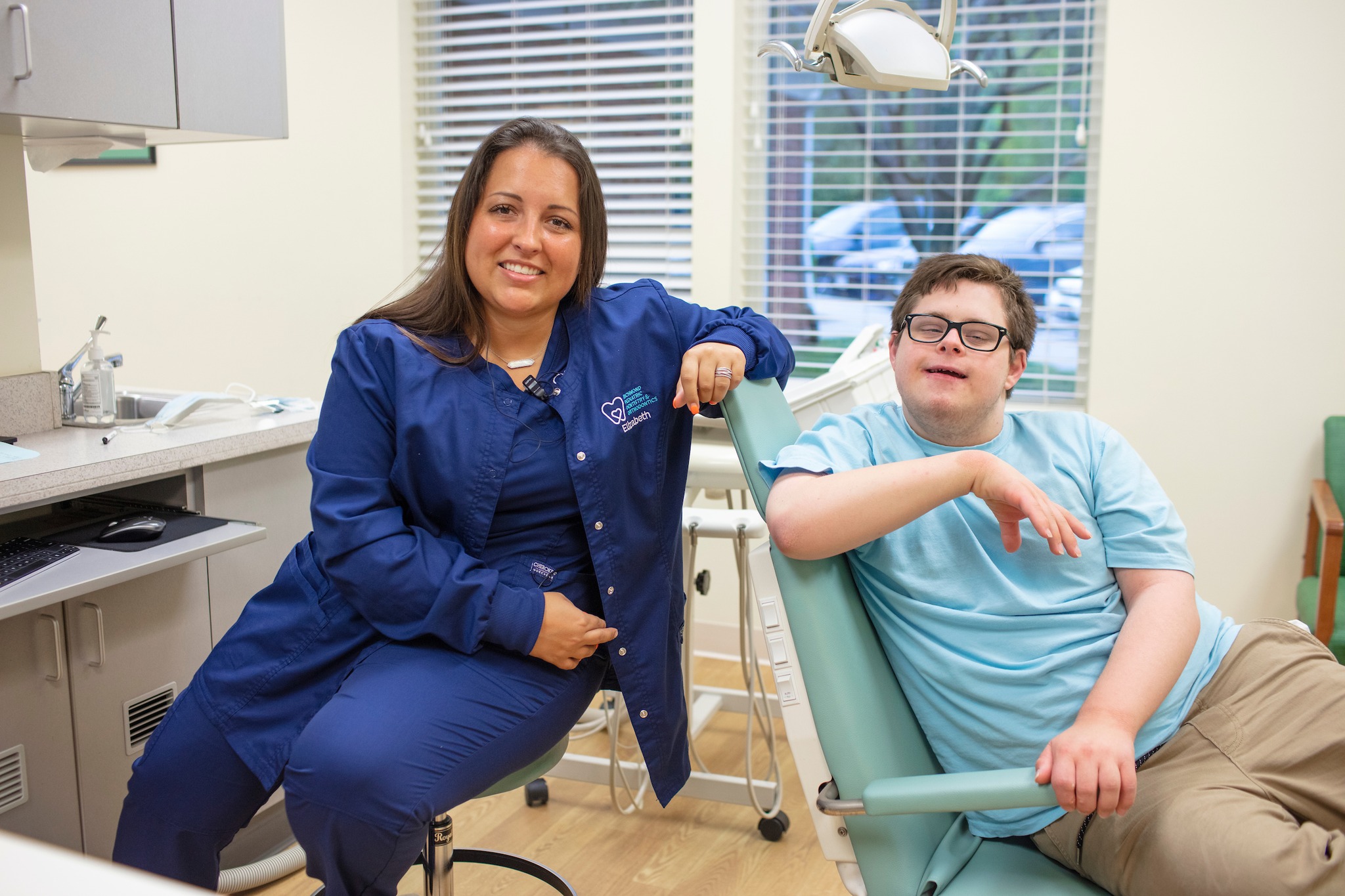 A smiling dental assistant named Elizabeth sitting beside a teenage patient with Down syndrome in a treatment chair at Richmond Pediatric Dentistry and Orthodontics, illustrating compassionate special needs pediatric dental care in Richmond, VA