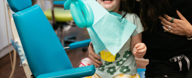 Smiling young girl holding a balloon animal in the dental chair beside a team member at Richmond Pediatric Dentistry and Orthodontics