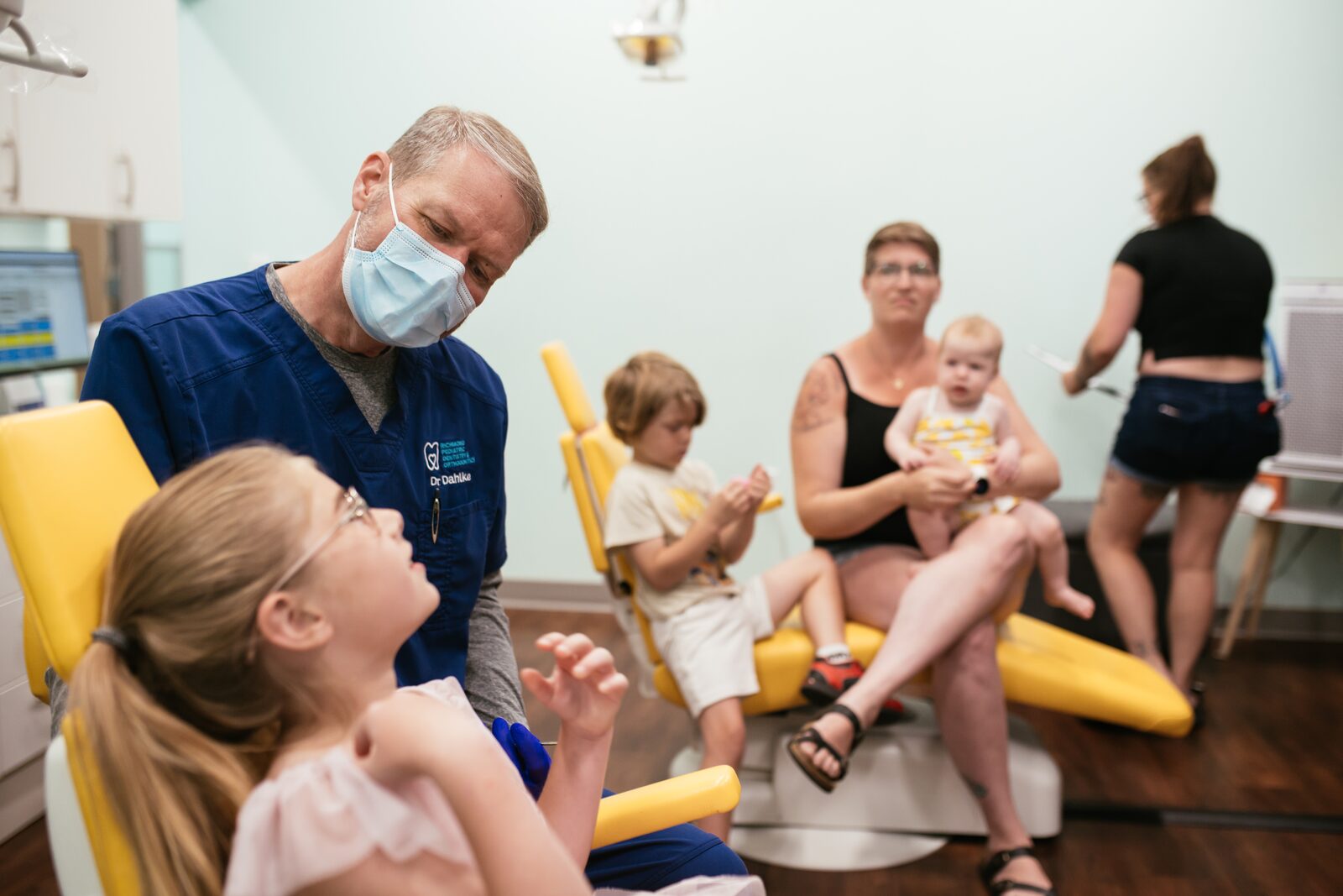 Your Child’s First Visit 6 Toddler and baby watching from a parent's lap while their big sister has a checkup with an RPDO pediatric dentist in Richmond, VA